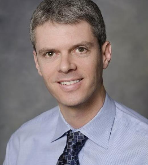 Photo of white man with dark gray hair on gray background wearing light blue shirt with dark blue tie with pattern